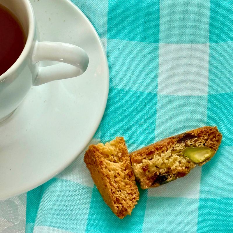 Cup of coffee with two cranberry pistachio biscotti pieces on a blue checkered tablecloth