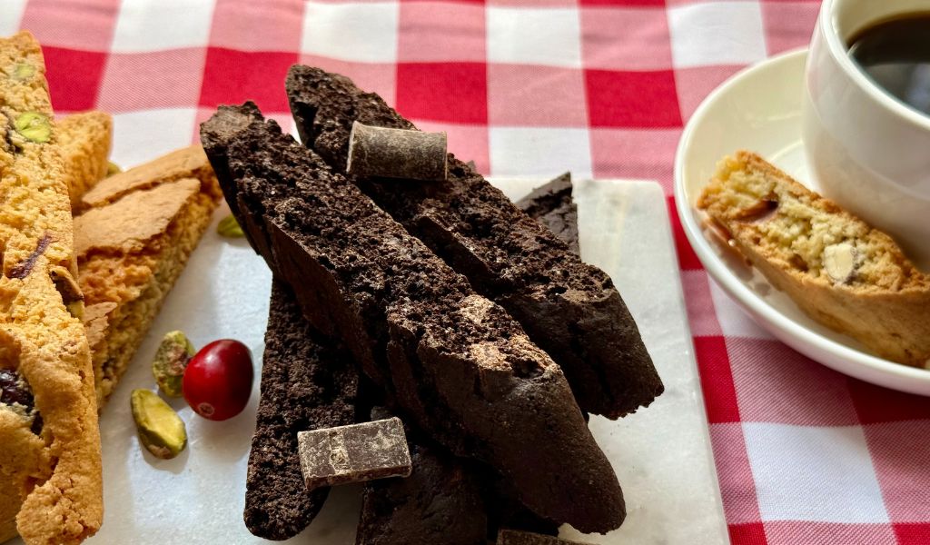Array of almond, cranberry pistachio, and double chocolate biscotti on a marble tray with a cup of coffee on a red checkered tablecloth.