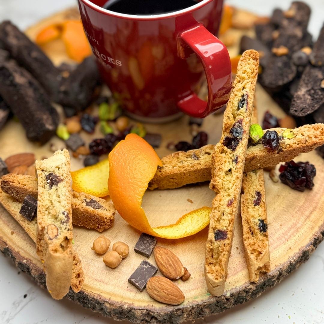 Overhead photo of assorted biscotti flavors arranged on a round wooden board with almonds, pistachios, orange rind, cranberries, caramel bits, and a cup of coffee in the center