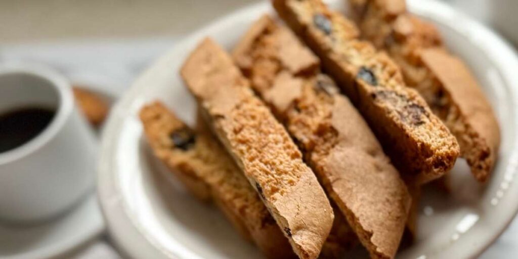 assortment of almond, cranberry pistachio, and double chocolate biscotti on a white ceramic plate beside espresso cups