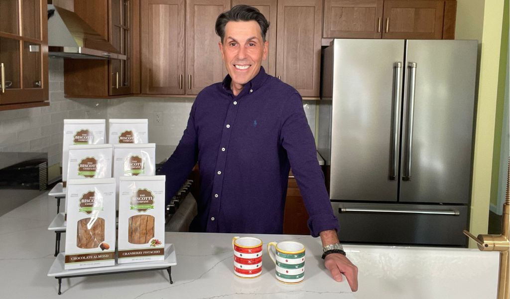 Bruno LoGreco standing beside a display of nine bags of gourmet biscotti with two cups of coffee on the table.