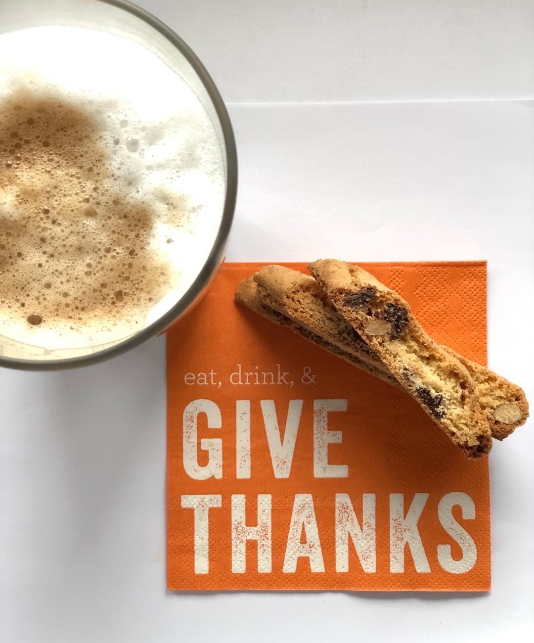 Tall latte with three chocolate almond biscotti fanned out on an orange napkin that reads Eat. Drink. Give Thanks.