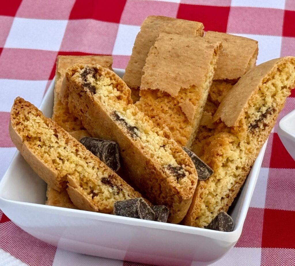 Chocolate Biscottini sitting in a white square plate with visible Belgian chocolate chunks and a few scattered chocolate pieces around them.