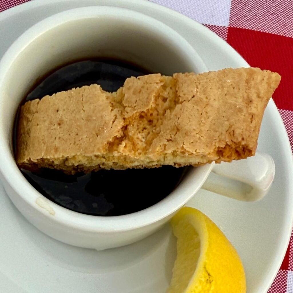 Lemon Biscottini being dunked into a cup of coffee, with a slice of lemon resting on the saucer to highlight the citrus flavor.