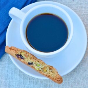 White espresso cup and saucer filled with coffee, with a Cranberry Pistachio Biscotti on the saucer