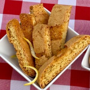 Lemon Biscottini in a white square dish with fresh lemon zest scattered around, set on a red checked tablecloth.