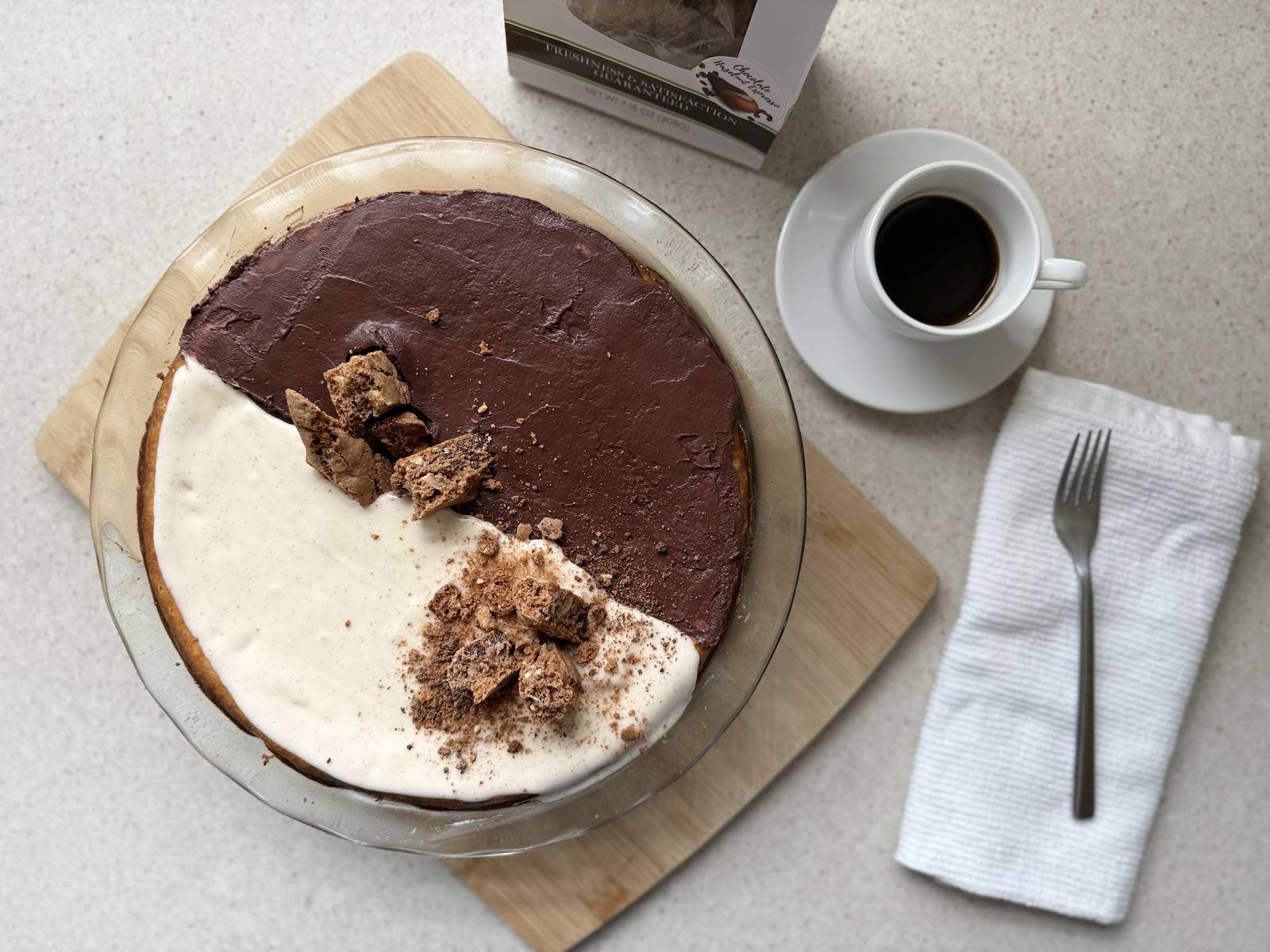 Overhead view of a whole biscotti cheesecake with black and white chocolate topping, Chocolate Hazelnut Espresso biscotti package, and an espresso cup