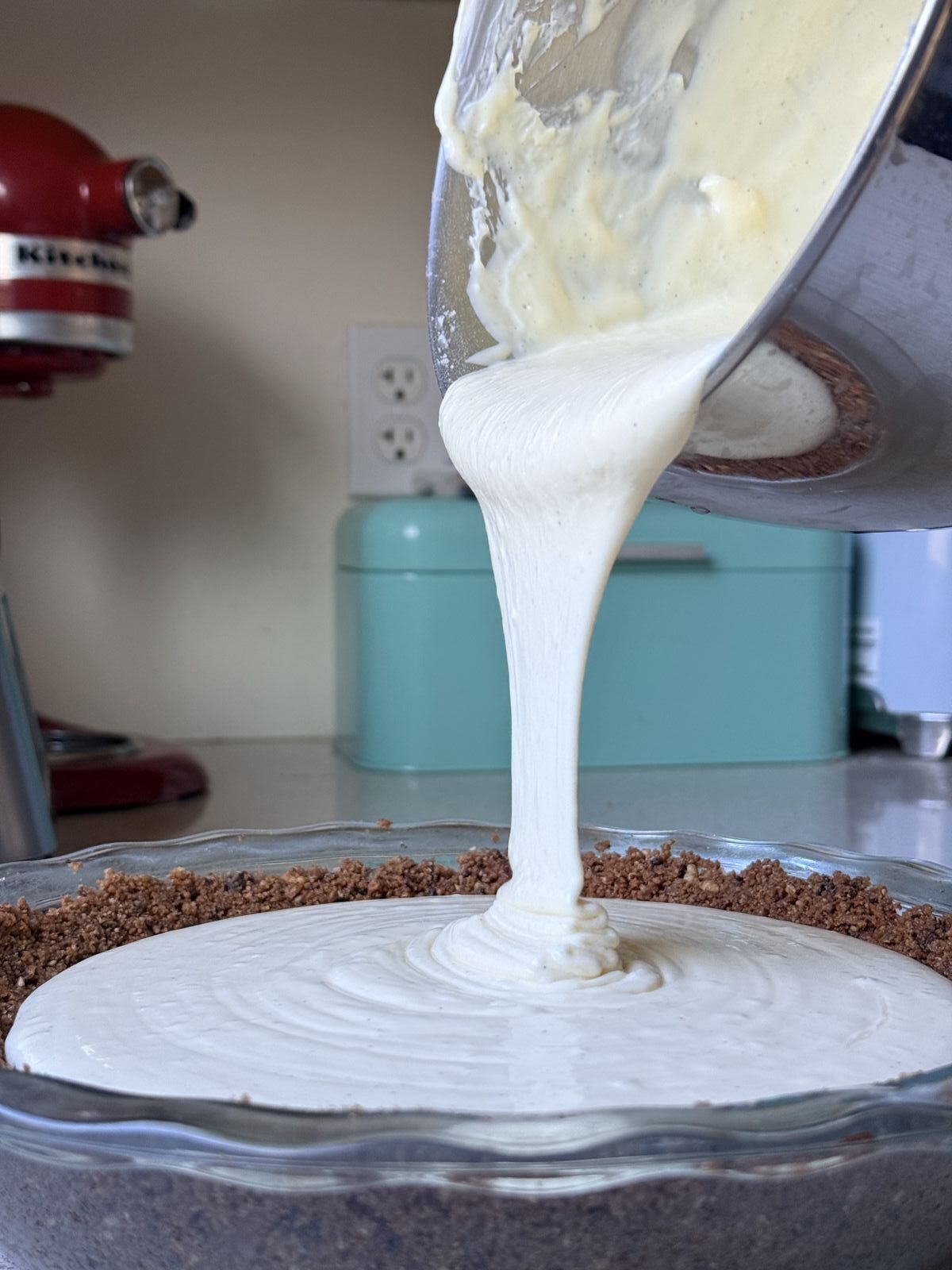 New York style cheesecake filling being poured from a mixer bowl into a Chocolate Hazelnut Espresso biscotti crust in a pie dish
