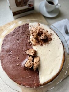 Two-tone chocolate and vanilla cheesecake half with cookie crumb topping on a glass plate, coffee cup in the background.