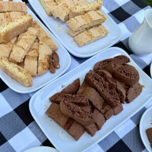 Three white plates with dessert slices: pale vanilla sponge bars, crumbly light cake pieces, and a plate of dense chocolate cake slices on a checkered tablecloth with a white pitcher nearby.